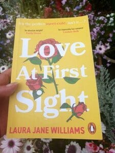 Paperback copy of Love At First Sight by Laura Jane Williams held in front of blooming garden flowers. The bright yellow cover features illustrated red roses and white text, with praise from Emily Henry and Lindsey Kelk above the title.