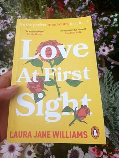 Paperback copy of Love At First Sight by Laura Jane Williams held in front of blooming garden flowers. The bright yellow cover features illustrated red roses and white text, with praise from Emily Henry and Lindsey Kelk above the title.