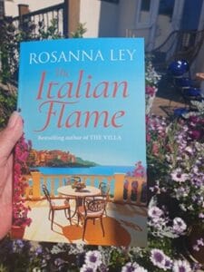 A hand holds a copy of The Italian Flame by Rosanna Ley against a vibrant garden background. The book cover shows a sun-drenched terrace with two chairs and a table overlooking a coastal Italian scene, with flowers and the sea in the distance. Purple and white daisies bloom around the foreground, adding a summery vibe.