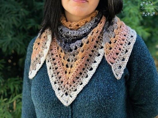 Triangle crochet shawl in warm gradient tones of cream, blush, copper, and charcoal, worn bandana-style around the neck over a dark teal sweater. The shawl features a classic granny stitch pattern with an open and textured design.