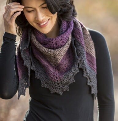 Woman wearing a cozy triangle crochet shawl in muted shades of plum, taupe, and charcoal, featuring a textured body and decorative picot edging. The shawl is wrapped snugly around her neck and shoulders over a long-sleeve black top.