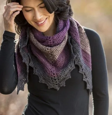 Woman wearing a cozy triangle crochet shawl in muted shades of plum, taupe, and charcoal, featuring a textured body and decorative picot edging. The shawl is wrapped snugly around her neck and shoulders over a long-sleeve black top.