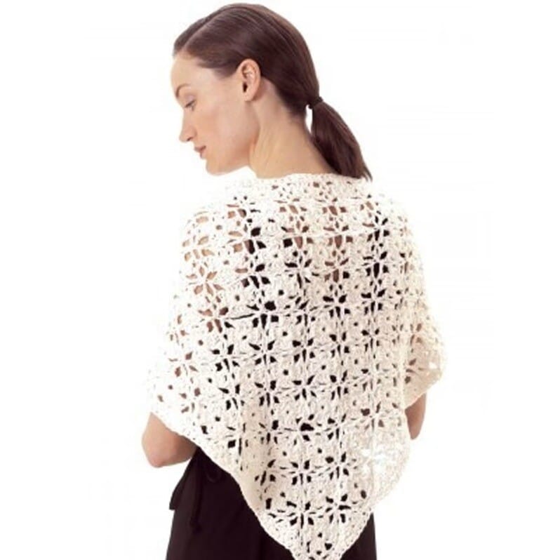 Woman wearing a cream-colored crochet shawl made from floral granny squares joined together, forming a triangular shape that drapes over her shoulders. The open, lacy design highlights the intricate motif work and lightweight texture.