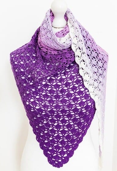 Triangle crochet shawl displayed on a mannequin, featuring a lacy shell stitch pattern and a gradient fade from deep purple to soft lavender and white. The elegant texture and smooth color transition create a delicate, feminine look.
