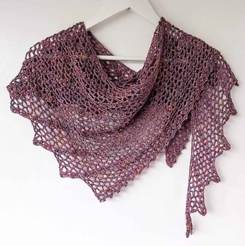 Lightweight crochet shawl in a dusty mauve yarn, featuring a lacy mesh pattern and gently scalloped edge. The asymmetrical triangle design is displayed on a white hanger against a plain wall, showcasing its drape and airy texture.
