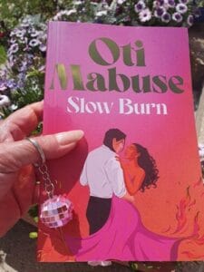 A hand holds a vibrant proof copy of "Slow Burn" by Oti Mabuse, featuring a romantic illustration of a couple dancing—her dress flaring into pink flames. The person’s hand holds a keyring with a small pink disco ball, and purple flowers bloom in the sunny background.