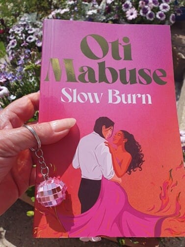 A hand holds a vibrant proof copy of "Slow Burn" by Oti Mabuse, featuring a romantic illustration of a couple dancing—her dress flaring into pink flames. The person’s hand holds a keyring with a small pink disco ball, and purple flowers bloom in the sunny background.