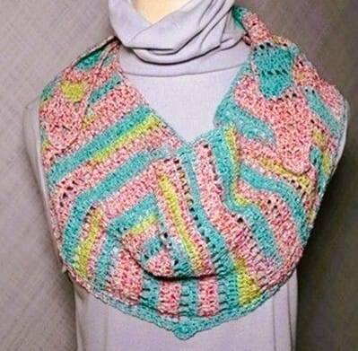 Bright and colorful crochet shawl in variegated yarn with stripes of pink, teal, yellow, and green, styled as a cowl on a mannequin dressed in a lavender turtleneck. The shawl features a chevron-like stitch pattern that adds texture and movement.