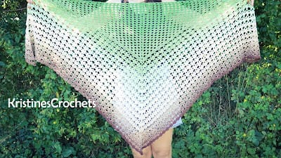 Large triangle crochet shawl held up outdoors in front of green foliage, featuring an airy mesh stitch and a color gradient from green to cream to dusty mauve.