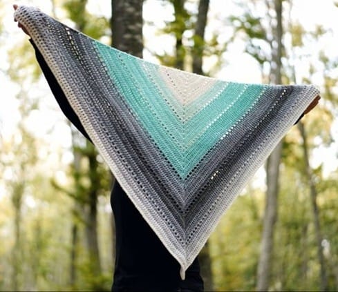Large triangle crochet shawl held up outdoors against a forest backdrop, featuring bold color blocking in cream, teal, gray, and black. The shawl’s rows of textured stitches and gradient stripes create a striking, modern design.