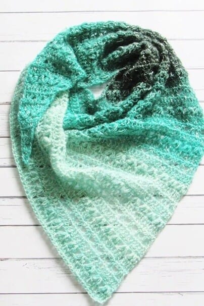 Triangle crochet shawl laid flat on a white wooden surface, featuring textured stitches and a smooth gradient from mint green to deep teal. The cozy, airy fabric makes it perfect for layering in cooler weather.