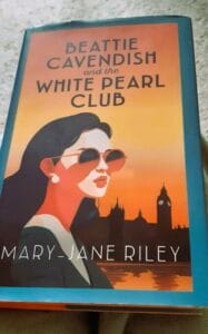 Cover of the book Beattie Cavendish and the White Pearl Club by Mary-Jane Riley. The image shows a stylish woman with dark hair, red lipstick, pearl earrings, and large sunglasses, set against an orange London skyline with Big Ben and the Thames at sunset. The design evokes a vintage, mysterious feel.