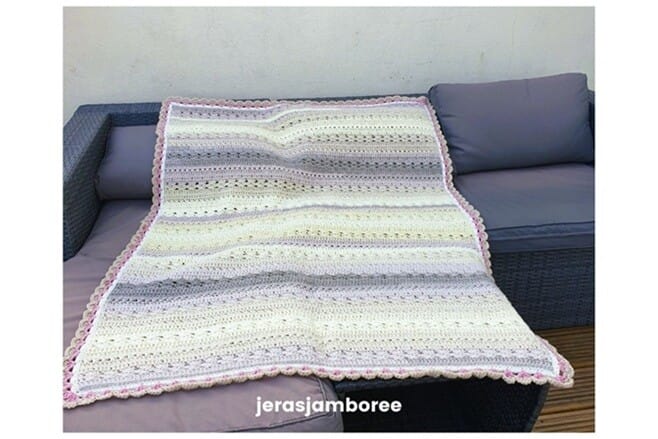 The image shows the Twilight Haven Crochet Blanket spread across a grey outdoor sofa. The blanket features soft pastel stripes in cream, yellow, grey, and lilac, finished with a pink scalloped border. The texture and stitch pattern are visible across the surface. The word “jerasjamboree” appears at the bottom of the image.