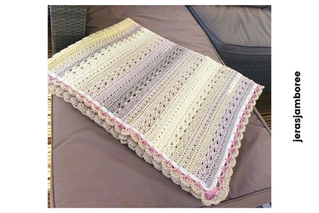 The image shows the Twilight Haven Crochet Blanket neatly folded and displayed on a grey outdoor sofa cushion. The blanket features pastel ombré stripes in cream, yellow, beige, and lilac, finished with a pink scalloped border. The layered stitches and textured pattern are visible along the folded edges. The word “jerasjamboree” is written vertically along the right-hand side of the image.