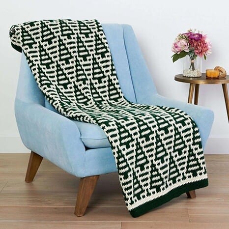 Overlay mosaic crochet blanket in dark green and cream with a bold repeating geometric pattern, draped over a light blue armchair beside a small table with flowers.