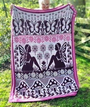 Overlay mosaic crochet blanket in white, pink, and dark brown, featuring large fairy silhouettes, butterflies, flowers, and mushroom motifs, held up outdoors in a garden.