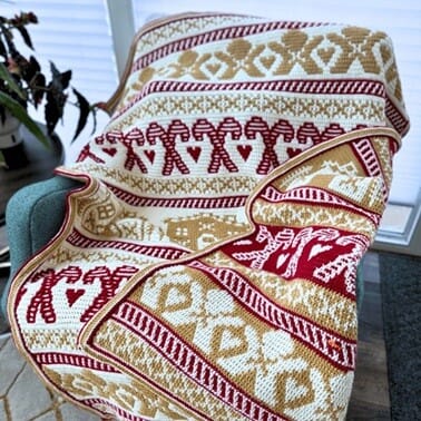 Overlay mosaic crochet blanket inspired by Christmas treats, in red, gold, and white, featuring motifs such as gingerbread people, hearts, houses, and snowflakes, draped over a chair.
