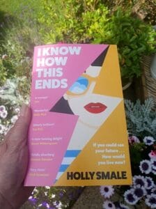 A hand holds a brightly colored copy of the book I Know How This Ends by Holly Smale, against a garden backdrop with purple and white flowers. The book cover features an illustrated woman's face with red lipstick and sunglasses, split diagonally with bold pink and yellow sections. Text on the cover includes praise quotes and the tagline, "If you could see your future… How would you live now?"
