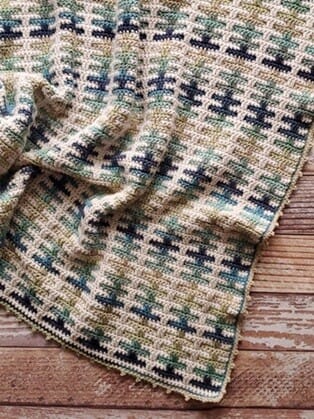 Inset mosaic crochet blanket in cream, green, and blue variegated yarn, featuring a repeating geometric pattern, laid out on a rustic wooden floor.