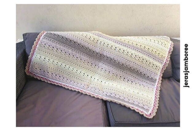 The image shows the Twilight Haven Crochet Blanket draped across the back and seat of a grey outdoor sofa. The blanket is displayed lengthwise, showing its pastel stripes in cream, yellow, lilac, and grey, with a pink scalloped border running along the edges. The stitch texture is clearly visible. The word “jerasjamboree” is written vertically along the right-hand side of the image.