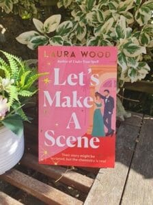 A paperback copy of Let’s Make a Scene by Laura Wood stands upright on a rustic wooden table outdoors. The book’s bright pink and red cover features an illustrated couple dressed for a glamorous night out. Surrounding the book are leafy variegated plants and a potted fern, with sunlight casting soft shadows across the scene.