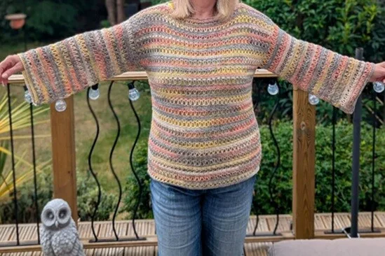 A woman wearing the Orla Crochet Pullover stands with arms outstretched on a wooden deck. The wide sleeves and relaxed fit of the sweater are clearly visible, along with the open stitch pattern and soft stripes in pastel tones. The photo is taken outdoors with a garden and decorative owl statue in the background.