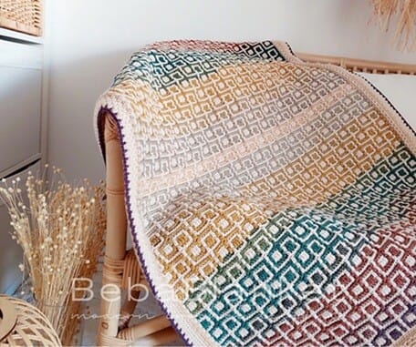 Overlay mosaic crochet blanket with diamond pattern in gradient colours of gold, rust, teal, and cream, draped over a wicker chair in a cosy indoor setting.