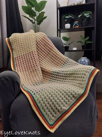 Inset mosaic crochet blanket with beige background and multicoloured squares in red, yellow, green, and blue, finished with a colourful striped border, draped over a dark grey armchair.