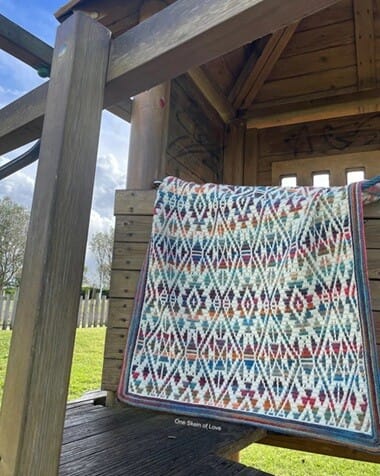 Overlay mosaic crochet blanket in cream, blue, red, and gold, featuring bold geometric tribal-inspired patterns, draped over a wooden playhouse outdoors.