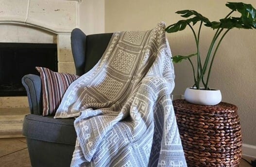 Tunisian mosaic crochet blanket in light grey and white, featuring different geometric square patterns, draped over a grey armchair beside a wicker side table with a potted plant.