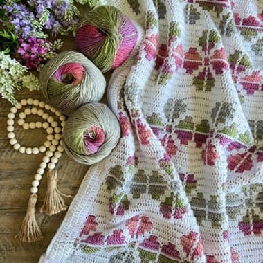 Overlay mosaic crochet blanket with pink, green, and purple floral motifs on a white background, displayed with coordinating yarn balls and flowers on a wooden surface.