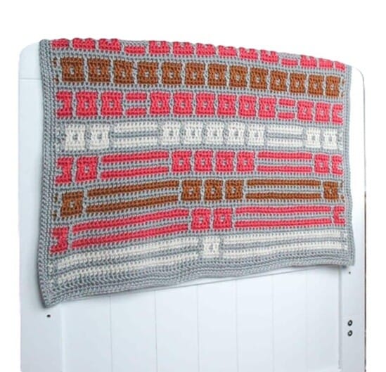 Inset mosaic crochet baby blanket in shades of grey, pink, brown, and white, featuring repeating geometric patterns, draped over a white wooden headboard.