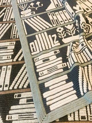 Overlay mosaic crochet blanket with a bookshelf design in cream, grey, and brown, featuring rows of books and decorative motifs, bordered in light blue.