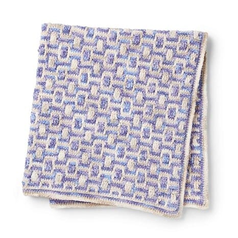 Overlay mosaic crochet blanket folded neatly, featuring a repeating geometric block pattern in lavender, cream, and light blue.
