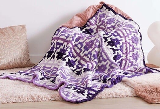 Overlay mosaic crochet blanket in shades of purple and white with geometric patterns, made in chunky yarn, draped over cushions on a soft rug.