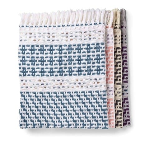 Overlay mosaic crochet blanket folded to show multiple panel designs in blue, white, peach, and purple, with fringe along the top edge.