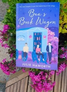 Paperback copy of Bea’s Book Wagon by Julie Haworth, with a bright illustrated cover showing a blue book wagon and three characters, surrounded by pink flowers outdoors.