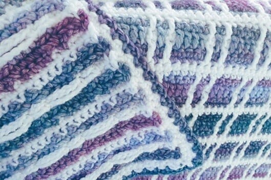 Close-up of the Fireside Plaid Crochet Blanket showing both the front and back: the tartan-style mosaic crochet on the right and the striped reverse on the left, in shades of purple, blue, and white.