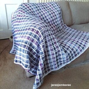 Fireside Plaid Crochet Blanket displayed on a sofa, showing its purple, blue, and white tartan design