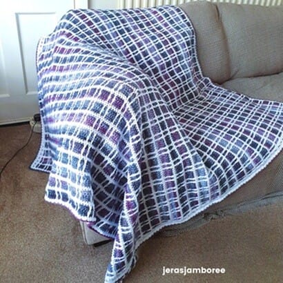 Fireside Plaid Crochet Blanket displayed on a sofa, showing its purple, blue, and white tartan design