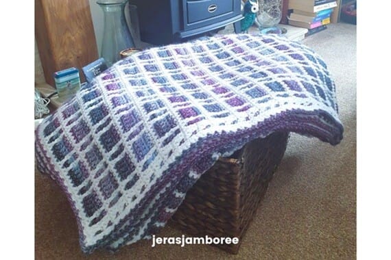 The Fireside Plaid Crochet Blanket draped over a woven basket beside a fireplace, showing its purple, blue, and white tartan design with a neat textured border, and the Jera’s Jamboree logo below.