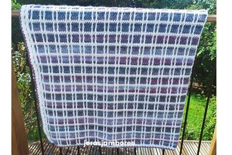 The Fireside Plaid Crochet Blanket hanging over a wooden railing outdoors, showing its purple, blue, and white tartan pattern created with mosaic crochet stitches, with the Jera’s Jamboree logo along the bottom.