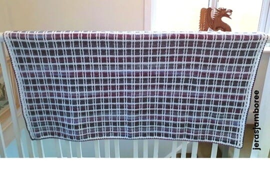 Fireside Plaid Crochet Blanket draped over an indoor railing, highlighting its purple, blue, and white tartan design with a neat border — a cosy couch throw and crochet blanket pattern from Jera’s Jamboree.