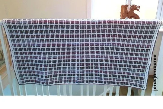 Fireside Plaid Crochet Blanket draped over an indoor railing, highlighting its purple, blue, and white tartan design with a neat border — a cosy couch throw and crochet blanket pattern from Jera’s Jamboree.