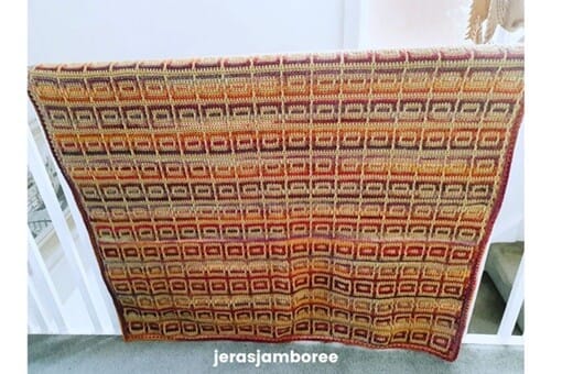 The Amberlight Crochet Blanket hanging over a white railing, showing the full inset mosaic pattern in warm amber, mustard, and plum tones with a neat border.
