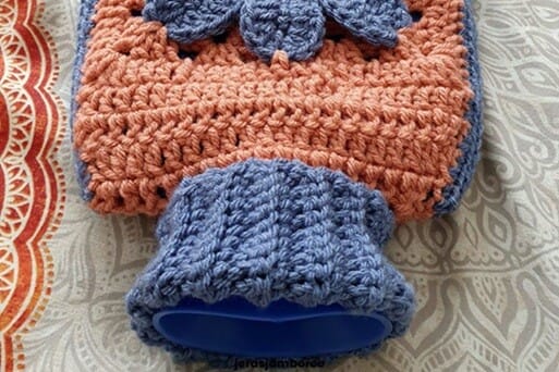 Close-up of the neck of the Cosmos Crochet Hot Water Bottle Cover showing the folded blue ribbed collar and the top of the orange crochet body with the blue flower detail visible above. A blue hot water bottle can be seen inside the cover.