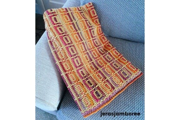 The Amberlight Crochet Blanket folded neatly on a light grey sofa, showing the inset mosaic stitch pattern in amber, orange, and purple tones.