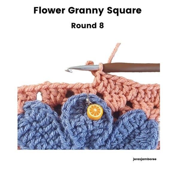 Flower Granny Square Round 8 showing the final round of orange double crochet stitches worked above the blue petals. A stitch marker shaped like an orange slice is attached to one petal, and a crochet hook is positioned in the working stitch.