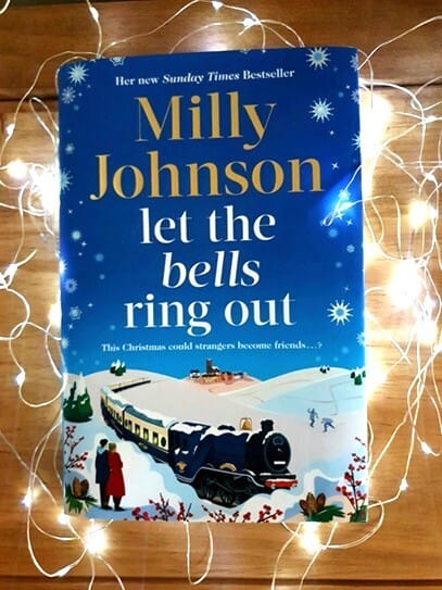 Hardback copy of Let the Bells Ring Out by Milly Johnson, a blue festive book cover featuring a steam train travelling through a snowy landscape with fairy lights glowing around it.
