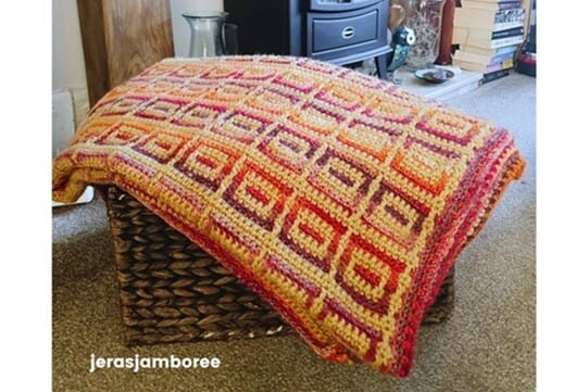 The Amberlight Crochet Blanket draped over a woven basket in front of a fireplace, showing the inset mosaic pattern in warm amber, orange, and purple tones with a textured border.